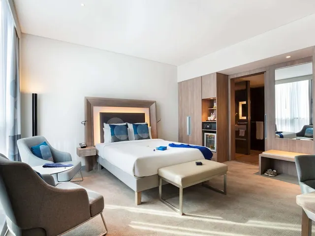 A hotel bedroom with a double bed, two armchairs, a bench at the foot of the bed, built-in wardrobes and a small fridge, with an open doorway to the bathroom.