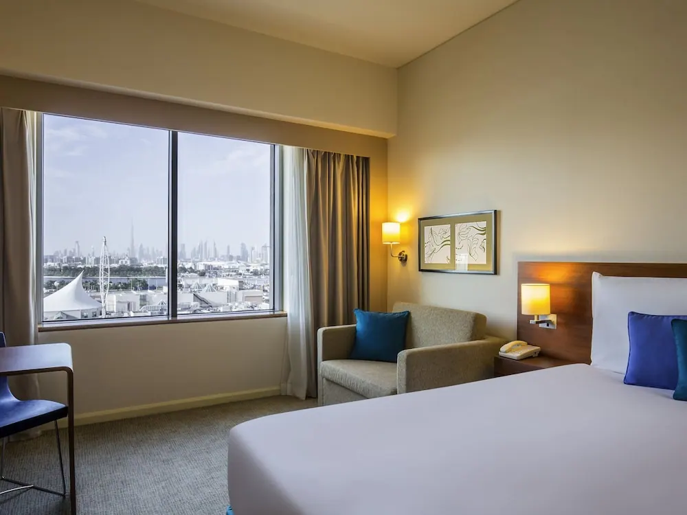 A hotel bedroom with a king bed, an armchair, a desk, and a window looking out over a city skyline with a distant tower.