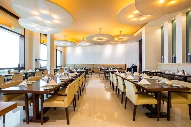 A formally set dining room with rows of wooden tables, cream upholstered chairs, banquette seating along the back wall and circular ceiling lights.