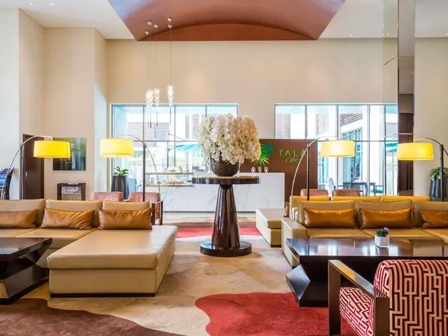 A lobby lounge with cream leather sofas, floor lamps, a white orchid arrangement on a dark pedestal table, and a TALA café counter in the background.