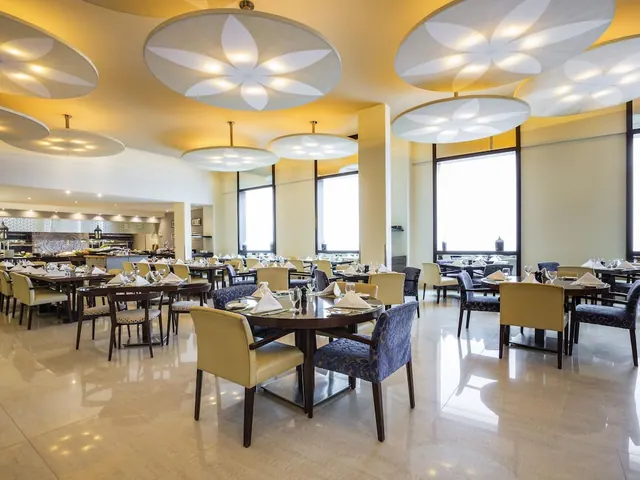 A restaurant dining room with round tables set with folded napkins and glassware, blue and yellow upholstered chairs, large flower-shaped ceiling lights, and a buffet station in the background.
