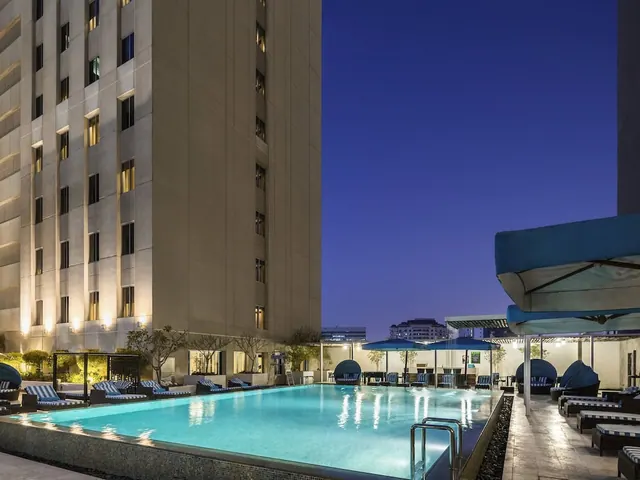 An outdoor pool at dusk with sun loungers and blue umbrellas beside a tall, lit hotel building under a deep blue sky.
