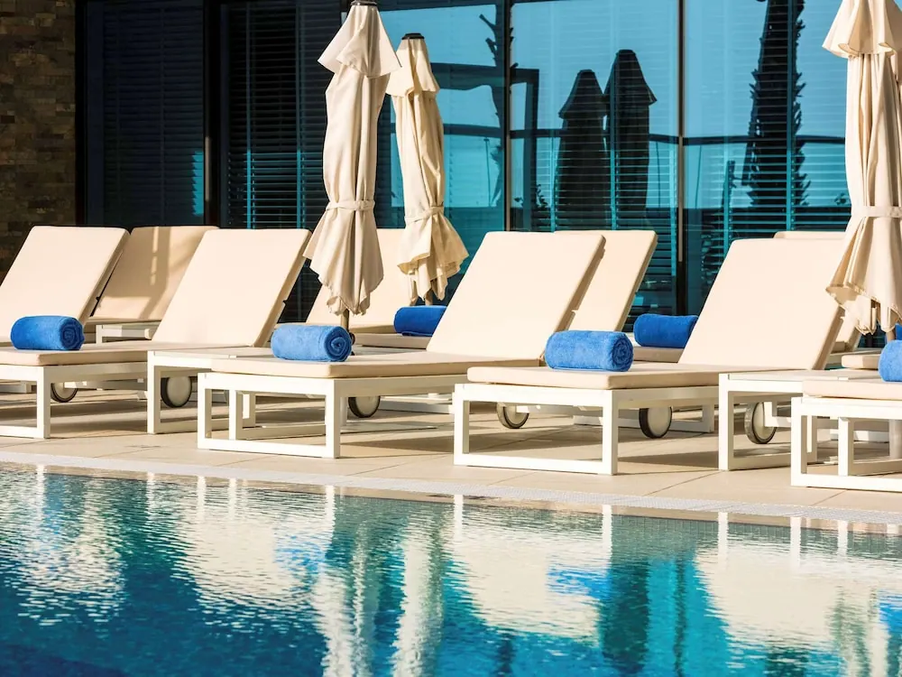 White sun loungers with cream cushions, blue rolled towels and closed cream umbrellas stand beside a swimming pool.