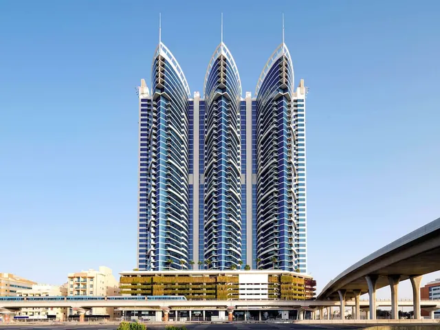Exterior view of a tall curved-glass tower with spired tops and a green-walled podium, with an elevated metro line and road in the foreground under a clear blue sky.