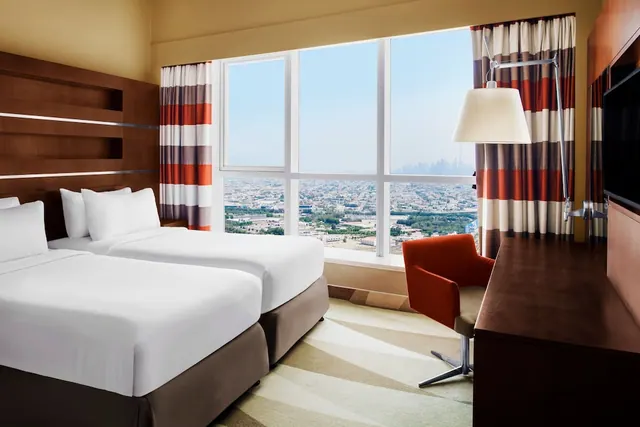 A bedroom with two single beds, an armchair, a wooden desk and television, with a large window looking out over a city.