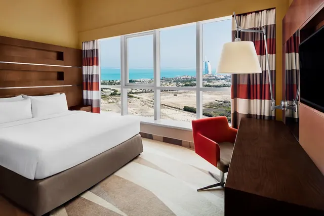 A bedroom with a king bed, a red armchair, a wall-mounted television and a large window looking out over the sea and the Burj Al Arab.
