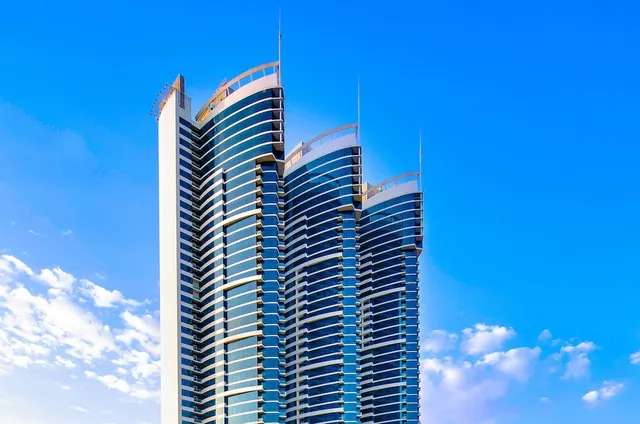 Low-angle view of a tall glass-fronted high-rise with curved balconies and rooftop railings against a blue sky with clouds.