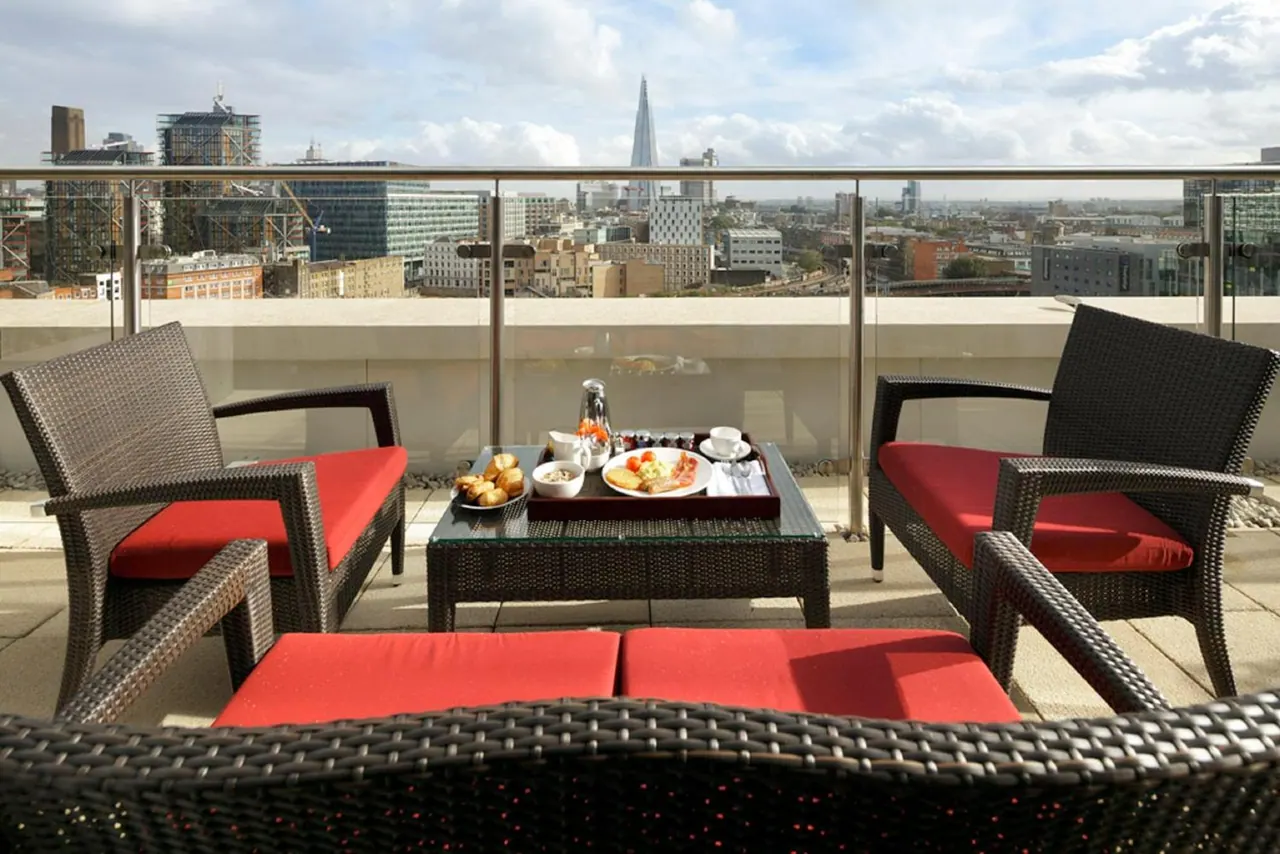 A furnished rooftop terrace with wicker armchairs, red cushions and a breakfast tray on a glass table, with a city view that includes The Shard.