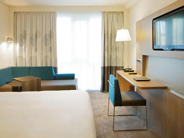 A hotel bedroom with a large bed with white bedding in the foreground, a teal sofa by the window, a wall-mounted television and a desk with a chair.