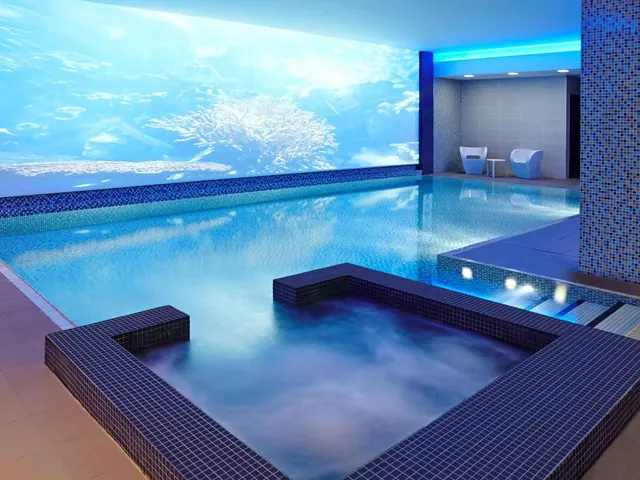 An indoor pool with a blue-lit printed wall mural and a mosaic-tiled hot tub in the foreground, with two chairs visible in the background.