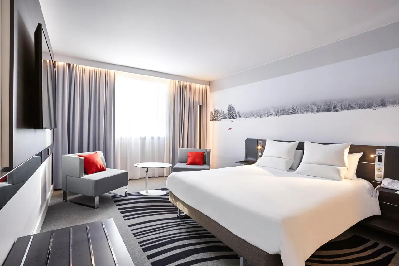 A bedroom with a made king bed, a snowy forest mural on the wall, two grey armchairs with red cushions, and a wall-mounted television.