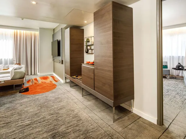 A hotel room with a bed, a wall-mounted television, a tall wooden storage unit and an orange rug, with an adjoining room visible through a doorway.