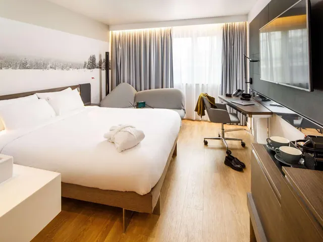 A hotel bedroom with a king bed, a grey sofa, a desk with a chair, a wall-mounted television and a coffee station on a wooden sideboard.