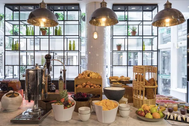 A breakfast buffet with pastries, fruit, juice glasses and cheese sits on a marble counter below pendant lamps and glass shelving.