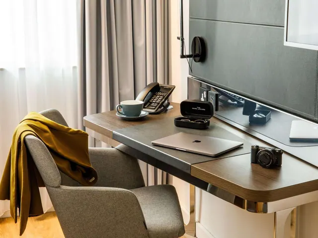 A desk holds a telephone, a coffee cup, a laptop, a camera and a sunglasses case beside an armchair with a mustard jacket draped over it, below a wall-mounted television.