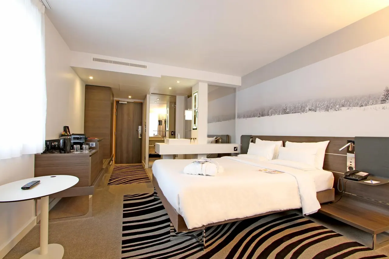 A hotel bedroom with a king bed, a folded bathrobe on the white bedding, a black and white snowy forest mural on the wall, and a bathroom visible through a glass partition.