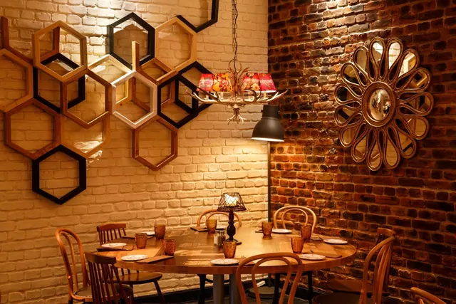 A round wooden dining table with bentwood chairs sits in a restaurant beside a whitewashed brick wall with hexagonal shelves and an exposed brick wall with a round mirror.