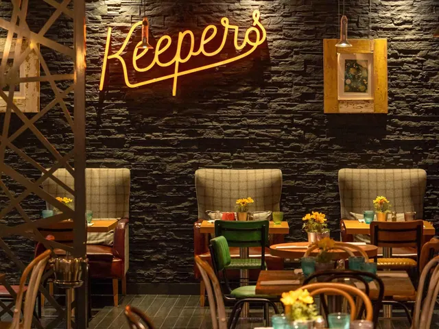 A dimly lit restaurant dining area with a glowing yellow neon Keepers sign on a dark brick wall, with upholstered booths and wooden tables.