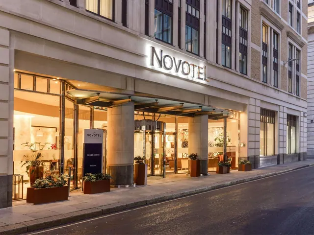 Exterior of the Novotel London Tower Bridge hotel entrance at dusk, with illuminated signage, a revolving door and potted plants along the pavement.