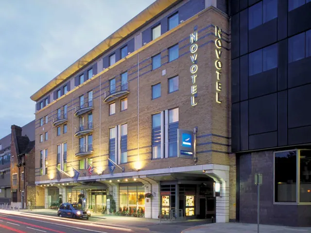 Exterior of a brick Novotel hotel building at dusk with illuminated vertical signs, a black cab on the street, and flags above the entrance.