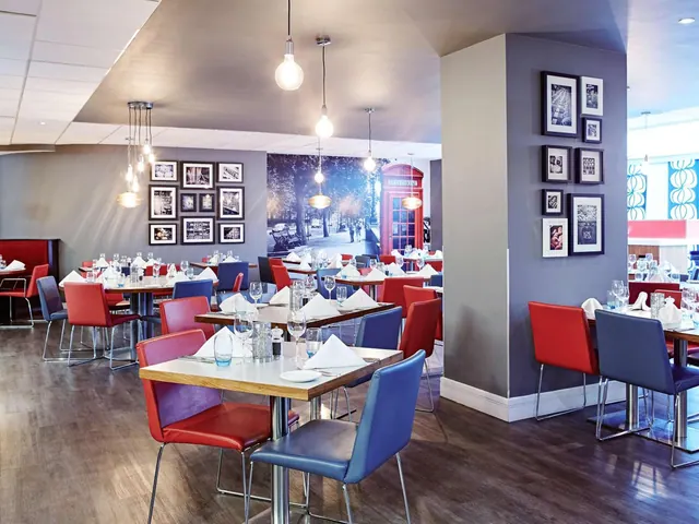 A restaurant dining room with red and blue chairs, tables set with folded napkins and glasses, pendant lights, and framed black and white photographs on the walls.