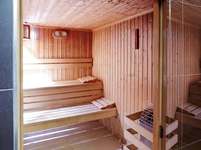 A wooden sauna cabin with slatted benches, a wall thermometer and hygrometer, and a heater with a metal guard, seen through a glass door.