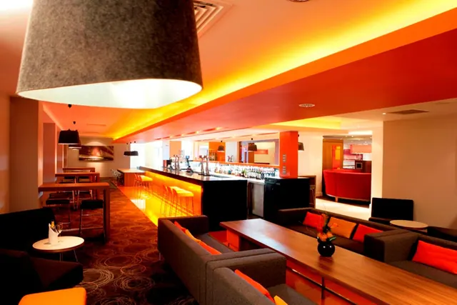 A hotel bar and lounge with orange ceiling lighting, a bar counter with stools, grey sofas with orange cushions and a wooden coffee table.