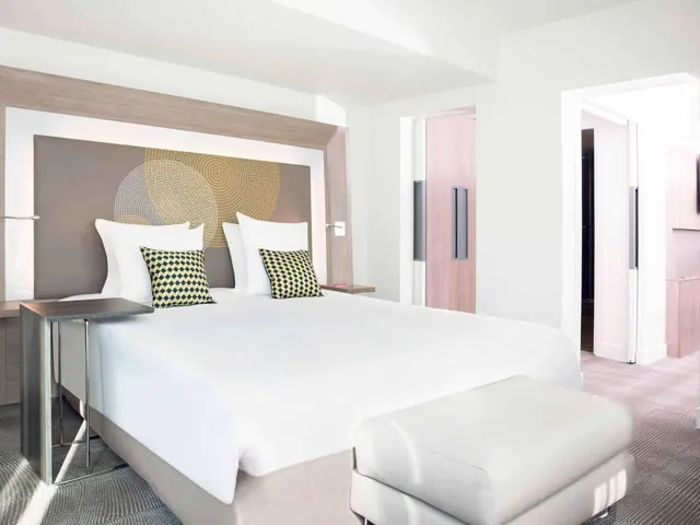 A bright modern hotel bedroom has a large double bed, a desk, storage cabinets and an upholstered seating area.