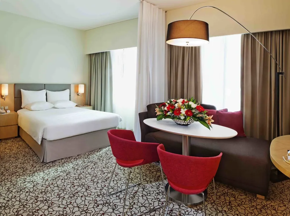 A hotel room with a made double bed, a grey sofa, a round white table holding a red flower arrangement, red chairs, and an arc floor lamp.