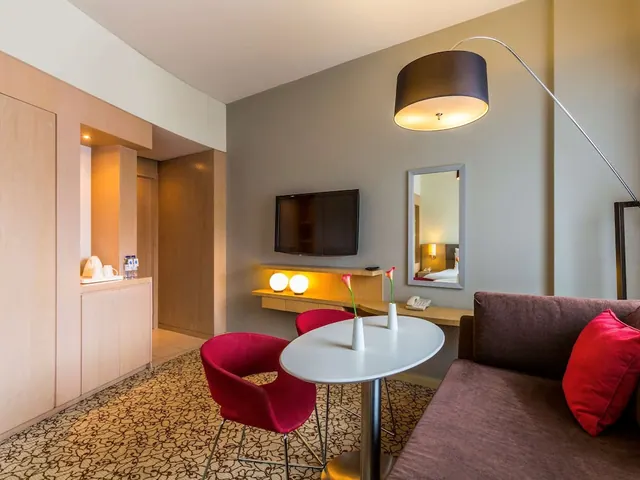 A hotel room living area with a round white table, two red chairs, a brown sofa with a red cushion, and a wall-mounted television.