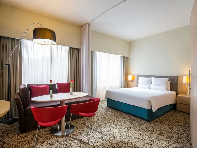 A hotel room with a double bed, a sofa and a round table with red chairs beside windows with sheer curtains.