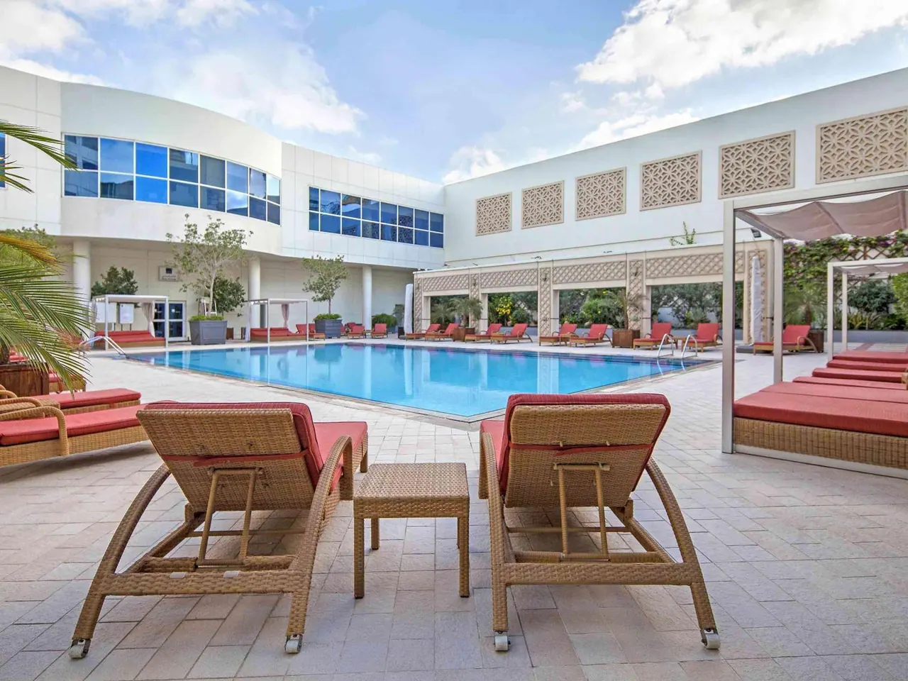 An outdoor pool in a courtyard with red sun loungers and wicker chairs on a tiled deck, surrounded by a white building with latticed panels.