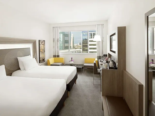 A hotel bedroom with two made beds, yellow armchairs, a desk with a kettle, and a window showing high-rise buildings.