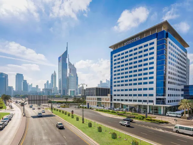 Exterior of a Novotel hotel tower beside a wide multi-lane road, with traffic and high-rise towers including the Emirates Towers visible in the background under a blue sky.