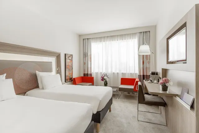 A hotel bedroom with two beds, two red armchairs, a desk with a coffee machine, and a wall-mounted television beside a window with sheer curtains.