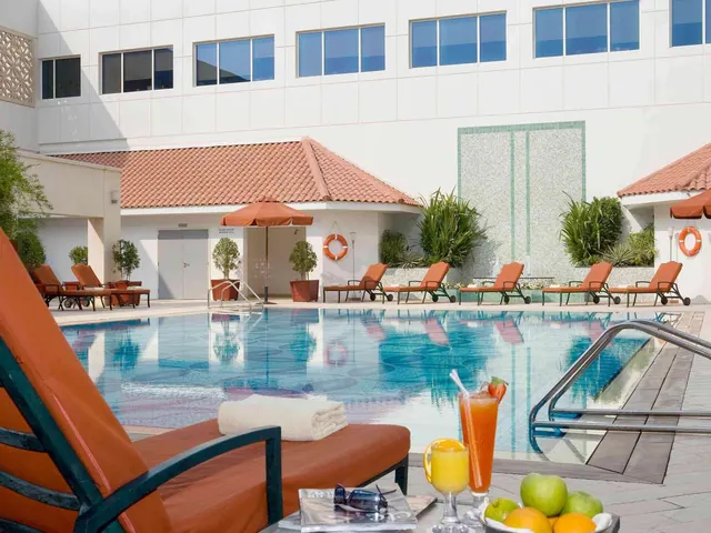 A large outdoor swimming pool with orange sun loungers and umbrellas beside a white hotel building, with a towel, drinks and fruit on a lounger in the foreground.