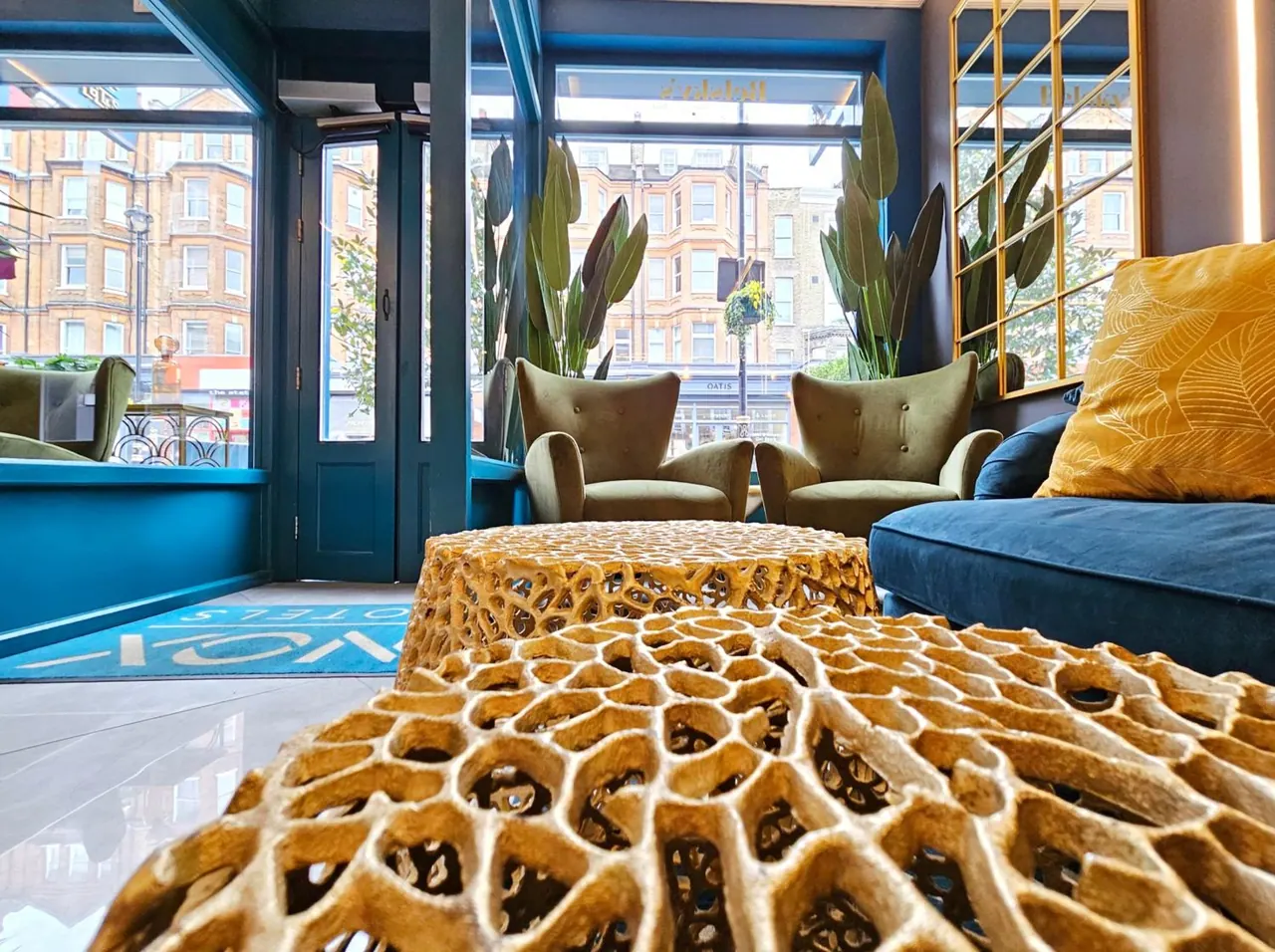 A lounge area with olive green armchairs, gold openwork coffee tables and a blue sofa, with windows looking onto a street of brick buildings.