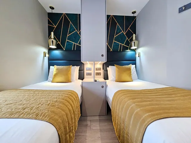 A bedroom with two twin beds topped with gold throws and cushions, a dark teal geometric feature wall, and a central cabinet with wall lights and sockets.
