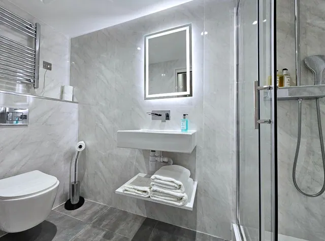 A bathroom with a wall-hung toilet, a wall-mounted basin with folded towels on a shelf below, a backlit mirror, and a glass-enclosed walk-in shower.