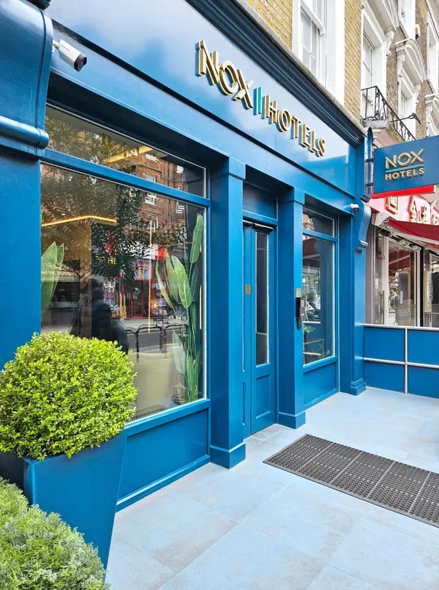The blue street-level facade of a Nox Hotels entrance with gold lettering, a glass door and window, a clipped topiary planter, and a doormat on pale paving.