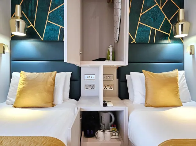 Two single beds with gold cushions and teal headboards sit either side of a central cabinet holding an iron, a kettle and tea and coffee items.