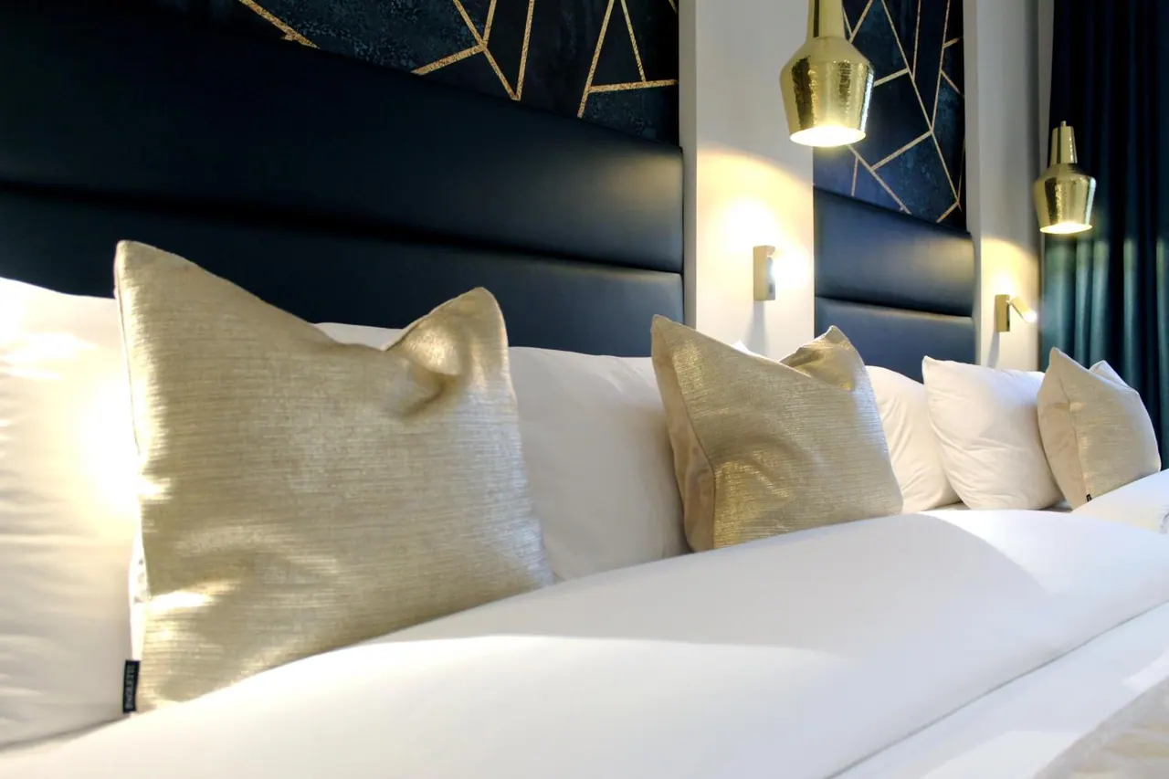 A made bed with white linen and gold cushions in front of a dark padded headboard, with gold pendant lamps and wall lights beside it.