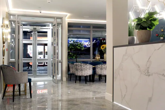 A lobby with a marble reception desk, glass entrance doors, upholstered chairs around a table, and potted plants, with a street visible through the windows.