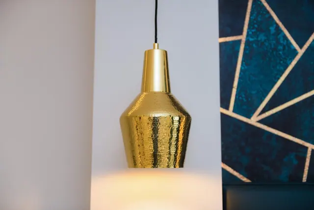 A hammered brass pendant lamp hangs in front of a white wall beside a navy wall panel with gold geometric lines.