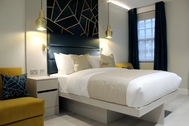 A bedroom with a double bed, a dark blue padded headboard, gold pendant lamps and a yellow armchair beside a window with navy curtains.