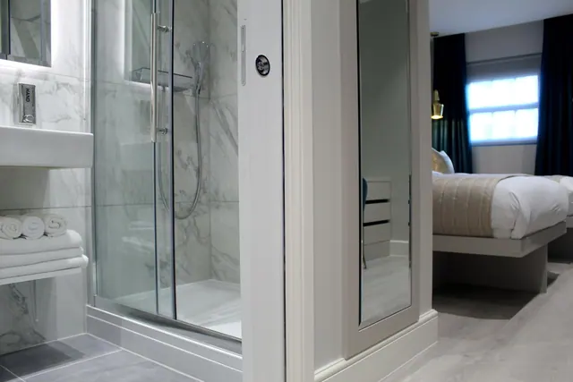 A glass-enclosed walk-in shower with marble-effect tiles sits beside a wall-mounted basin and folded white towels, with a bed visible through the open doorway.