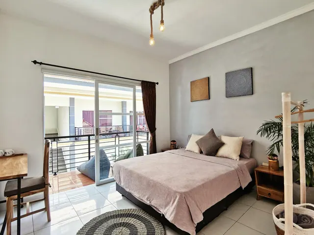 A bedroom with a double bed, a wooden desk and chair, and a sliding glass door opening onto a balcony with a bean bag chair.