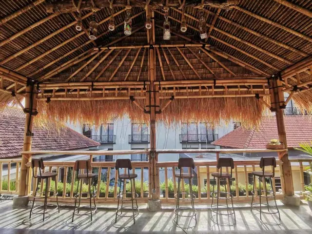 An open-air thatched bamboo pavilion with six bar stools along a bamboo railing, with neighbouring white buildings and red tile roofs visible behind.