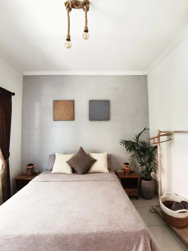 A bedroom with a double bed dressed in mauve linen and cushions, two wall pictures, a rope pendant light, bedside tables with lamps, and a potted palm.