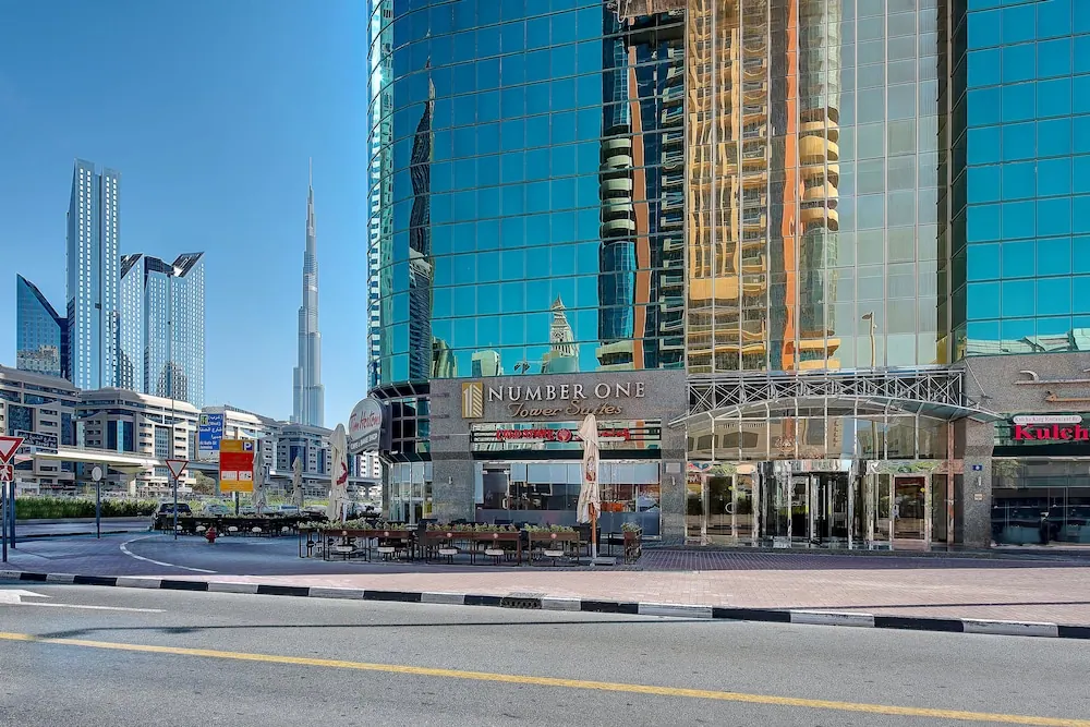 Exterior of the Number One Tower Suites building with its reflective glass facade, street-level entrance, outdoor tables with umbrellas, and the Burj Khalifa visible in the background.